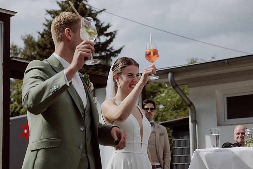 Während ihrer Hochzeitsfeier im Freien im Fischerstübchen Hemmingen erheben Braut und Bräutigam ihre Gläser zu einem fröhlichen Toast. Der Bräutigam trägt einen grünen Anzug, während die Braut in ihrem weißen Kleid mit Schleier glänzt. In der Nähe mischen sich die Gäste untereinander und ein Tisch mit Gläsern steht für weitere Feierlichkeiten bereit.