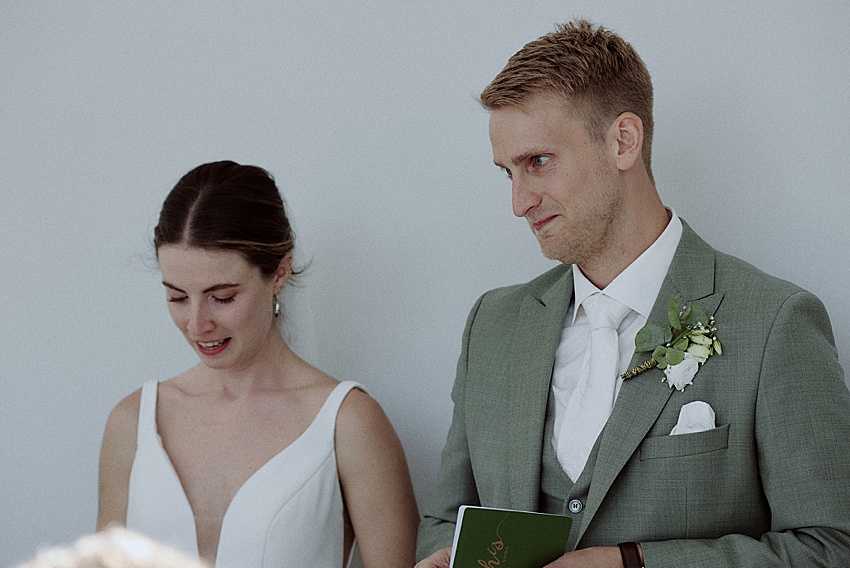 Eine Braut im weißen Kleid und ein Bräutigam im hellgrauen Anzug stehen zusammen bei ihrer Hochzeitsfeier. Der Bräutigam hält ein Büchlein in der Hand und beide wirken emotional. Sie befinden sich in einem gut beleuchteten Innenraum mit schlichtem Hintergrund, möglicherweise im malerischen Fischerstübchen Hemmingen.