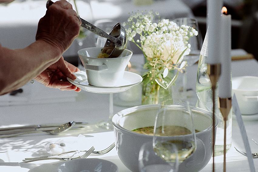 Bei einer Hochzeitsfeier im Fischerstübchen Hemmingen schöpft eine Person anmutig Suppe in eine weiße Schüssel an einem elegant gedeckten Tisch. Der Tisch ist mit weißen Tellern, Weingläsern, einem Blumengesteck mit zarten weißen Blumen und einer großen weißen Kerze ausgestattet.