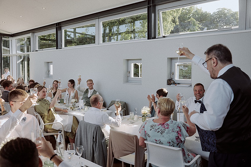 Bei einer Hochzeitsfeier im Fischerstübchen Hemmingen erheben die Gäste an einem formellen Esstisch ihre Gläser zum Toast. Der gut beleuchtete Raum verfügt über große Fenster, viele Gäste sind elegant gekleidet und schöne Blumen schmücken die Tische.