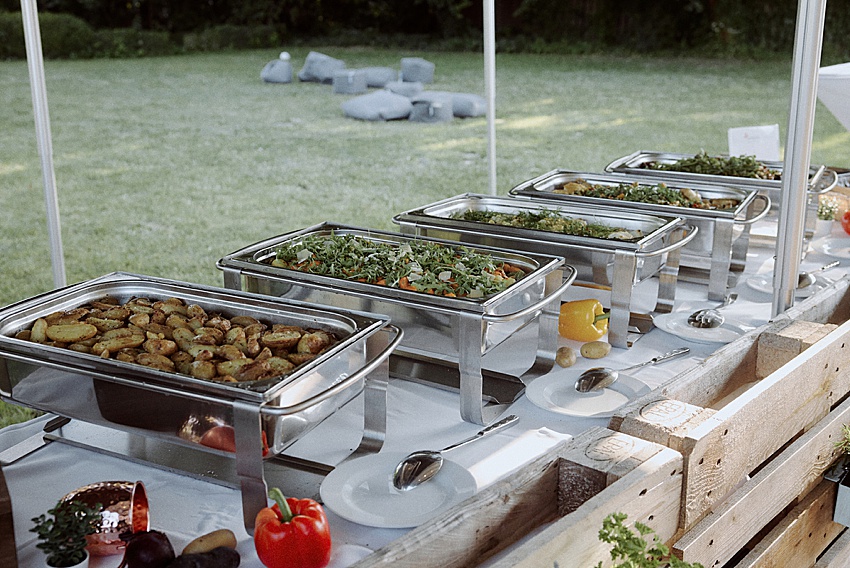 Bei der Hochzeitsfeier vor dem Fischerstübchen Hemmingen schmückt ein Buffettisch die Rasenfläche und bietet Köstlichkeiten wie Bratkartoffeln und frische Salate. Mit bereitliegenden Tellern und Besteck genießen die Gäste dieses Festmahl unter einem Baldachin, umgeben von der natürlichen Kulisse aus Bäumen und Gras.