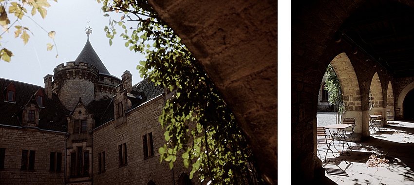 Ein Steingebäude mit Turm und steilem Dach, teilweise von grünen Weinreben bedeckt, erinnert an die Erhabenheit von Schloss Marienburg. Ein gewölbter Gang mit Steinsäulen führt zu einem schattigen Bereich mit Metallstühlen und -tischen. Diese malerische Kulisse ist perfekt für ein Hochzeitsshooting, da das Sonnenlicht bezaubernde Schatten auf den Boden wirft.