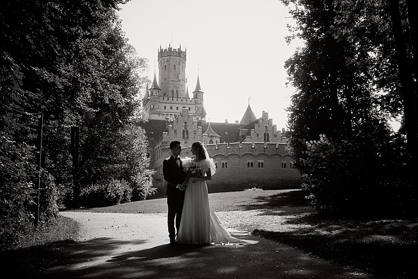 Braut und Bräutigam stehen auf einem Weg vor Schloss Marienburg, umgeben von Bäumen. Dieses Hochzeitsshooting fängt sie in Schwarzweiß ein, einander gegenüberstehend, während das Sonnenlicht Schatten auf den historischen Boden wirft.