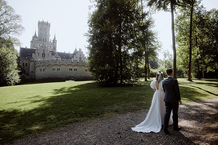 Braut und Bräutigam stehen vor Schloss Marienburg, einem historischen Schloss, umgeben von üppigen Bäumen und einem gepflegten Rasen. Die Sonne wirft Schatten auf die Grasfläche unter dem klaren Himmel. Perfekt für ein Hochzeitsshooting trägt die Braut ein weißes Kleid und eine weiße Schleife im Haar, während der Bräutigam einen Anzug trägt.