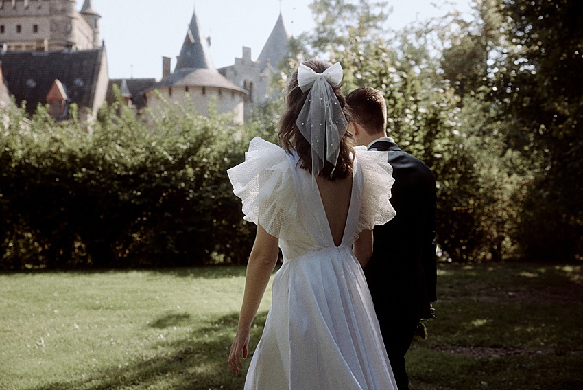 Braut und Bräutigam schlendern in Richtung einer üppigen, grünen Landschaft am Schloss Marienburg, einer exquisiten Kulisse für ihr Hochzeitsshooting. Die Braut in ihrem weißen Kleid mit Puffärmeln und der Bräutigam in seinem dunklen Anzug werden vom historischen Charme des Schlosses umrahmt.