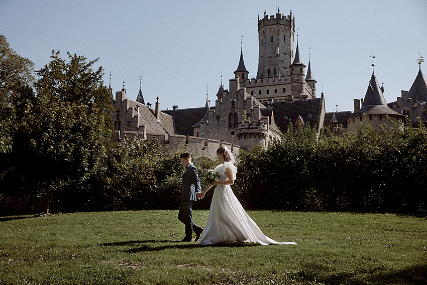 Braut und Bräutigam gehen Hand in Hand über eine Wiese, perfekt für ein Hochzeitsshooting. Die Braut trägt ein weißes Kleid, der Bräutigam einen blauen Anzug. Hinter ihnen steht das prächtige Schloss Marienburg, ein exquisiter historischer Ort mit seinen Türmen und Spitzen, die in den klaren Himmel ragen.