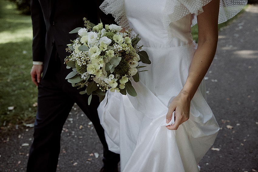 Eine Person in einem weißen Kleid mit Rüschenärmeln hält während eines Hochzeitsshootings einen Strauß weißer und grüner Blumen in der Hand und geht auf einem Weg. Neben ihr läuft eine weitere Person in dunkler Kleidung. Der malerische Hintergrund zeigt die Grasfläche und Bäume in der Nähe von Schloss Marienburg.