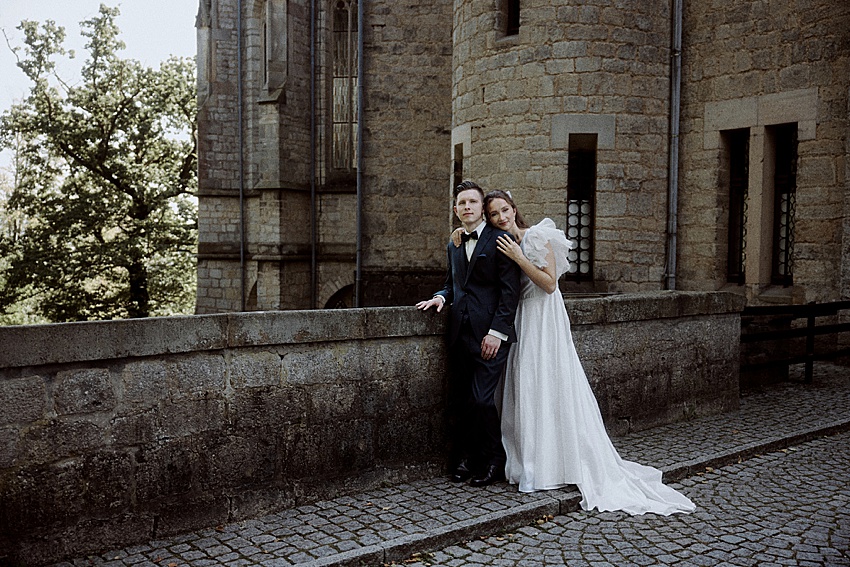 Braut und Bräutigam stehen auf einem Kopfsteinpflasterweg neben einem Steingebäude im Schloss Marienburg. Die Braut in ihrem langen weißen Kleid legt ihre Hand für ihr Hochzeitsshooting leicht auf die Schulter des Bräutigams. Dieser historische Ort verleiht ihrem besonderen Moment zeitlosen Charme.