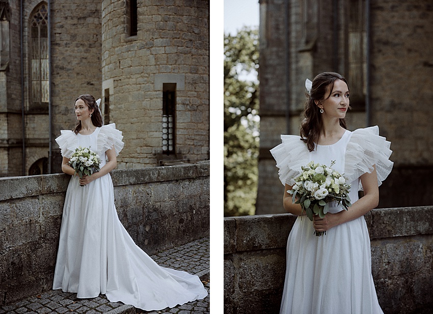 An einem historischen Ort steht eine Frau in einem weißen Brautkleid mit Rüschenärmeln an einer Steinmauer und hält einen Blumenstrauß in der Hand. Die Kulisse dieses Hochzeitsshootings besteht aus einem alten Steingebäude und üppigen Bäumen, die den zeitlosen Charme von Schloss Marienburg unterstreichen.