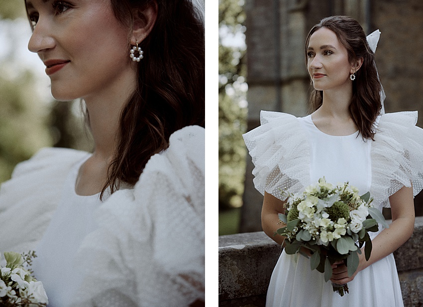 Eine Frau in einem weißen Kleid mit Rüschenärmeln hält einen Blumenstrauß. Sie steht in der Nähe von Schloss Marienburg, einem historischen Steinbau. Ihr langes dunkles Haar ist mit einem weißen Band geschmückt und sie trägt Perlenohrringe, die die Essenz eines bezaubernden Hochzeitsshootings einfangen.