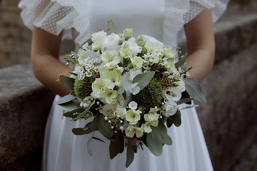 Eine Person in einem weißen Kleid hält einen Blumenstrauß, perfekt für ein Hochzeitsshooting. Der Strauß enthält grüne und weiße Blüten mit Grün. Vor der Kulisse der Steinmauer von Schloss Marienburg verleiht er diesem historischen Ort einen Hauch von Eleganz.