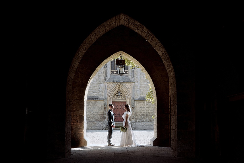 Braut und Bräutigam stehen sich unter einem Torbogen im Schloss Marienburg gegenüber. Der Steinbogen umrahmt sie im Tageslicht und schafft einen perfekten Moment für ein Hochzeitsshooting. Die Braut hält einen Blumenstrauß und beide sind formell gekleidet. Im Hintergrund ist eine verzierte Tür des historischen Veranstaltungsortes zu sehen.