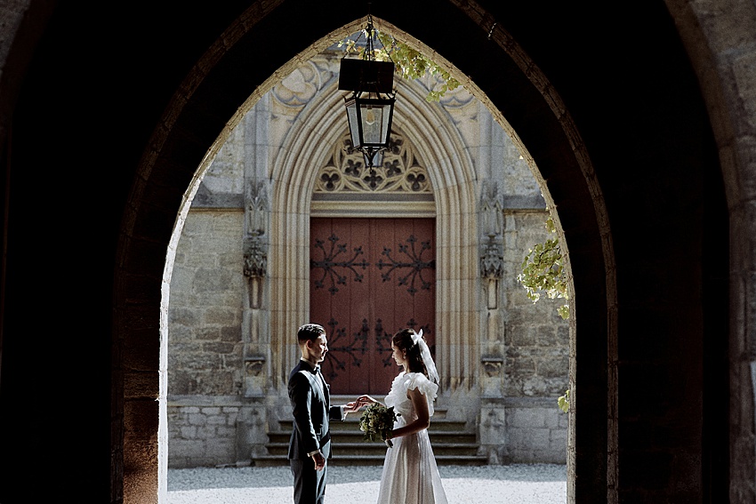 Ein Paar steht sich unter einem Torbogen im Schloss Marienburg, einem historischen Ort, gegenüber. Die Frau trägt ein weißes Kleid und hält einen Blumenstrauß, während der Mann einen dunklen Anzug trägt. Kunstvolles Mauerwerk und eine geschlossene, dekorative Holztür bieten eine atemberaubende Kulisse für ihr Hochzeitsshooting.