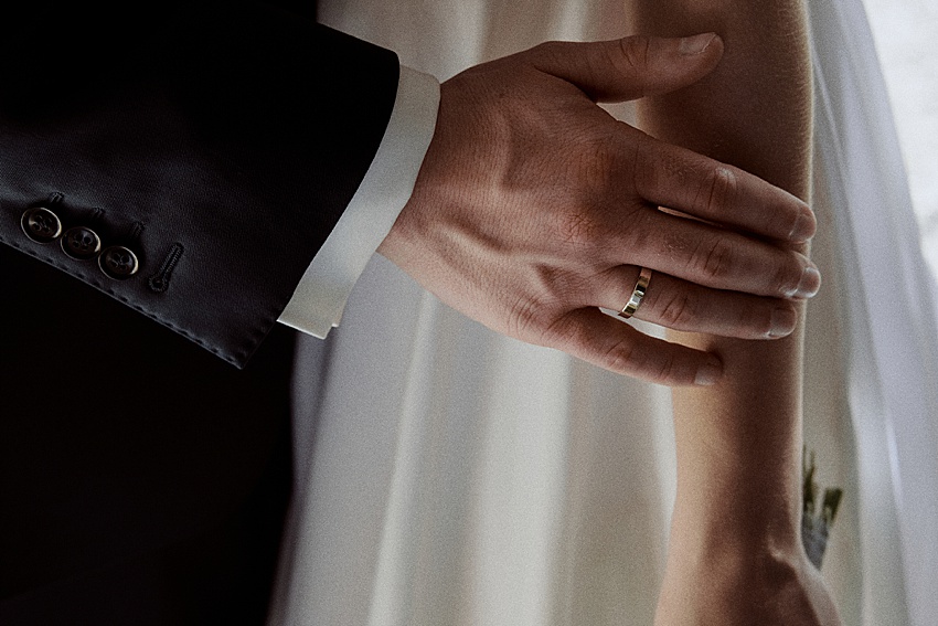 Nahaufnahme der linken Hand einer Person, die einen goldenen Ring trägt und auf dem Arm einer anderen Person in einem weißen Kleid ruht. Die Szene vor der Kulisse von Schloss Marienburg zeigt ein elegantes Hochzeitsshooting mit dem Ringträger in einem dunklen Anzug mit sichtbaren Ärmelknöpfen.