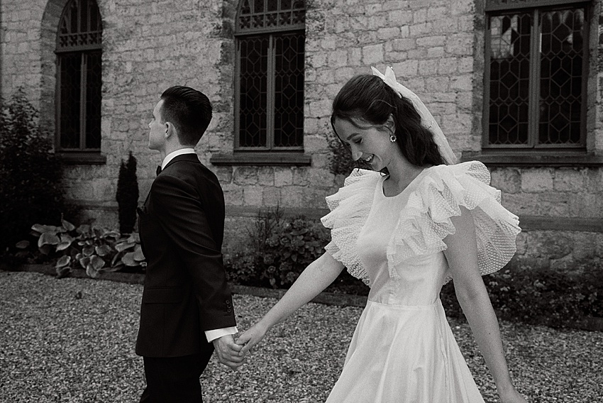 Eine Braut in einem weißen Kleid mit Rüschenärmeln und Schleier hält während ihres Hochzeitsshootings die Hand eines Bräutigams im dunklen Anzug. Sie gehen auf einem Kiesweg vor Schloss Marienburg, dessen Steinfassade und Bogenfenster den Moment einrahmen. Die Braut lächelt und der Bräutigam blickt weg.