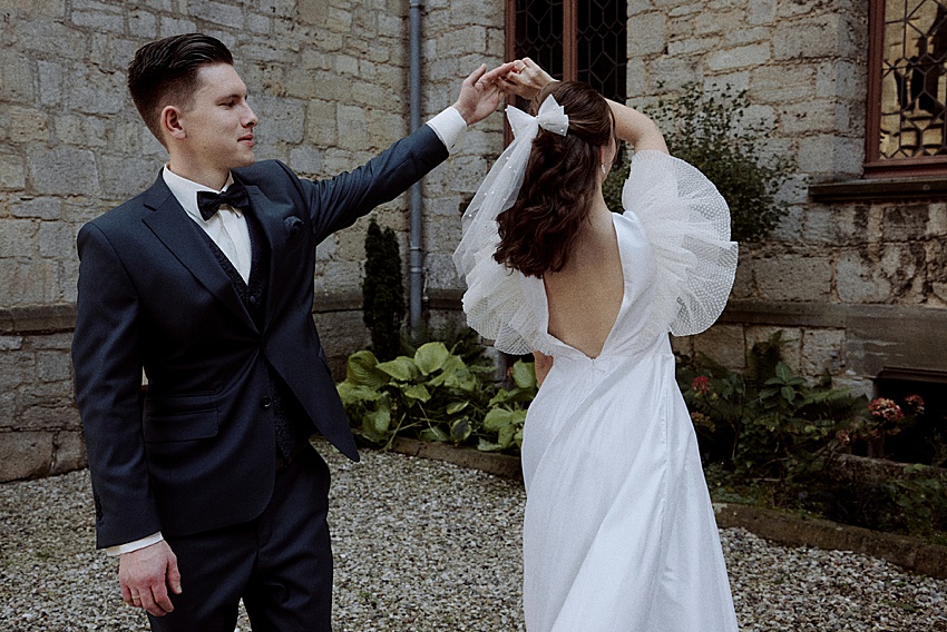 Ein Bräutigam im dunklen Anzug wirbelt eine Braut in einem weißen Kleid mit voluminösen Ärmeln und offenem Rücken herum. Sie stehen draußen auf Kies am historischen Ort von Schloss Marienburg, mit Pflanzen in der Nähe, die die perfekte Kulisse für ihr Hochzeitsshooting bieten.