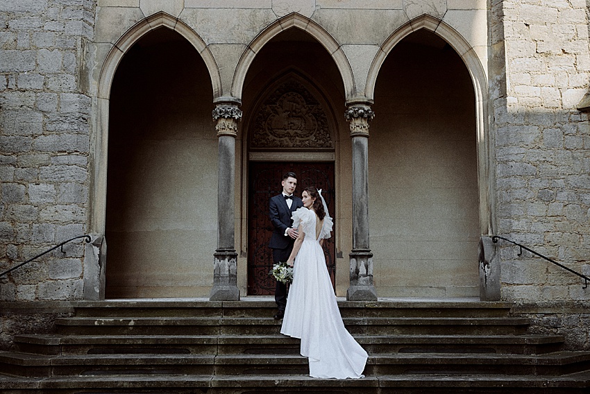 Braut und Bräutigam stehen auf Steinstufen vor Schloss Marienburg, einem alten Gebäude mit drei Torbögen. Die Braut in einem weißen Kleid mit einem Blumenstrauß in der Hand und der Bräutigam in einem dunklen Anzug sorgen mit geschnitzten Details über dem Mittelbogen für einen atemberaubenden Hochzeitsshooting-Moment.