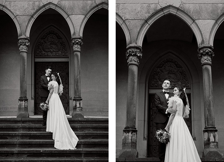 Ein Schwarzweißfoto zeigt ein Paar im Hochzeitskleid auf den Stufen von Schloss Marienburg. Die Braut in einem langen Kleid hält einen Blumenstrauß, während der Bräutigam neben ihr steht. Sie posieren unter einem mit kunstvollen Verzierungen verzierten Torbogen an diesem historischen Ort.