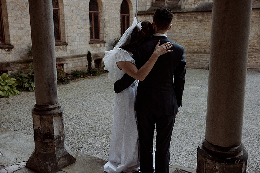 Eine Braut in weißem Kleid und Schleier hat ihren Arm um einen Bräutigam im dunklen Anzug gelegt. Sie stehen draußen, mit dem Rücken zur Kamera, im Hof von Schloss Marienburg – einem wahrhaft historischen Ort, perfekt für ihr Hochzeitsshooting zwischen Steinmauern und Säulen.