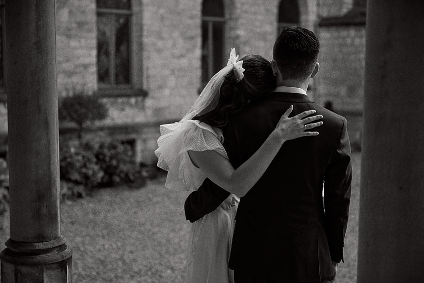 Ein Schwarz-Weiß-Foto zeigt ein umarmtes Paar vor dem historischen Schloss Marienburg. Die Frau, in Kleid und Schleier, legt sanft ihren Arm auf die Schulter des Mannes. Er steht im Anzug, mit dem Rücken zur Kamera. Diese zauberhafte Hochzeitsshooting-Kulisse weckt zeitlose Romantik im Freien.