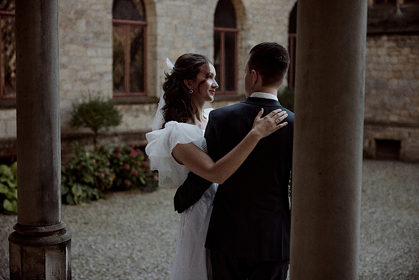 Ein Paar steht eng beieinander im Schloss Marienburg, einer bezaubernden Außenkulisse mit Steinmauern. Die Frau im weißen Kleid legt sanft ihre Hand auf die Schulter des Mannes, während sie sich unterhalten. Die perfekte Kulisse für ein Hochzeitsshooting, er ergänzt ihre Eleganz in einem dunklen Anzug.