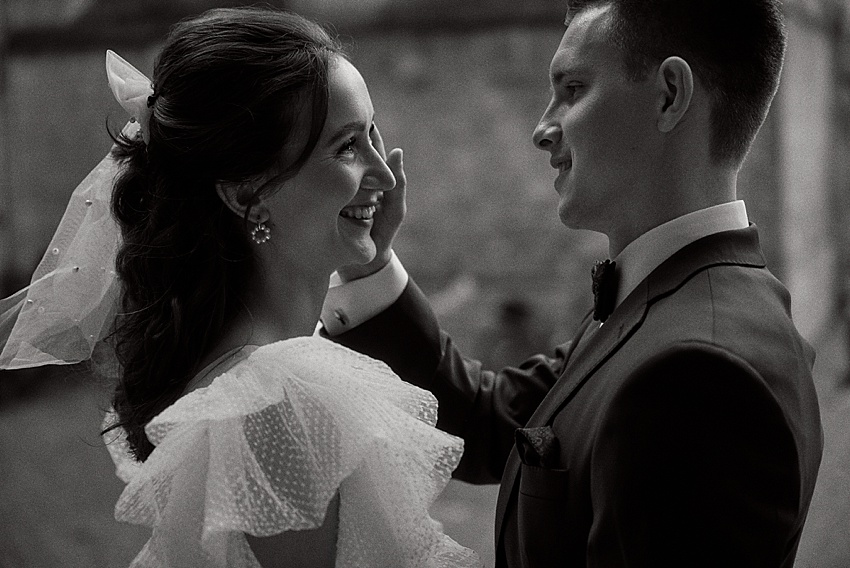 Ein Schwarzweißfoto eines Paares an seinem Hochzeitstag auf Schloss Marienburg. Die Braut lächelt, trägt ein Kleid mit Rüschen an den Schultern und einen Schleier, während der Bräutigam im Anzug sanft ihr Gesicht berührt. Sie sind im Freien mit einem unscharfen Hintergrund.