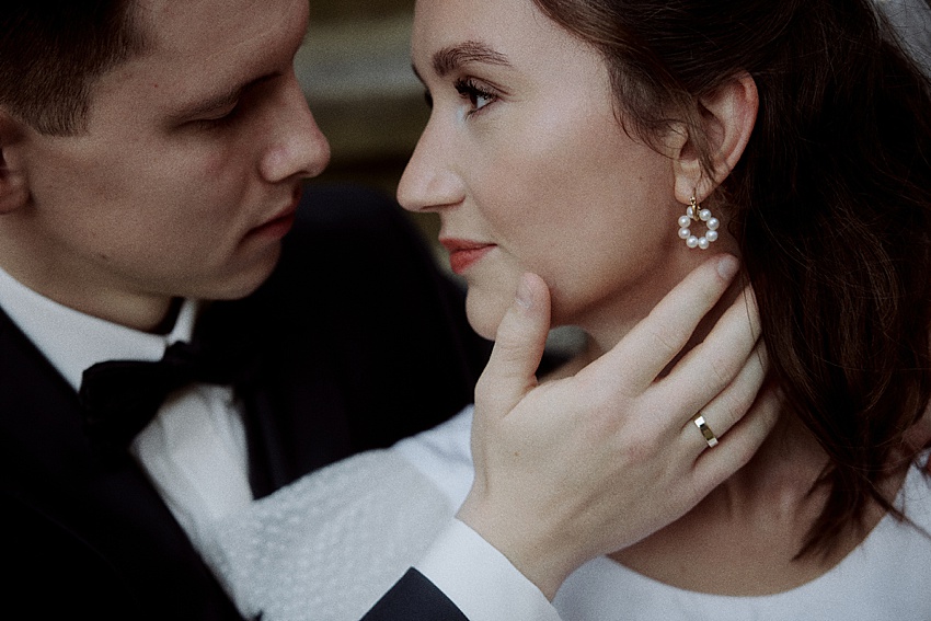 Ein formell gekleidetes Paar sitzt eng beieinander und genießt einen zärtlichen Moment während des Hochzeitsshootings auf Schloss Marienburg. Der Mann, der eine schwarze Fliege trägt, hält sanft das Kinn der Frau. Sie blickt aus dem Bild, geschmückt mit Perlenohrringen und weißem Kleid, und fängt die Eleganz dieses historischen Ortes ein.